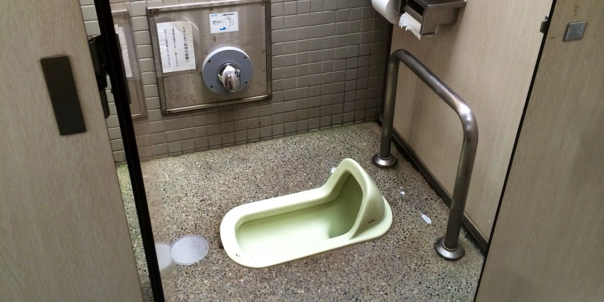 Japanese squat toilet in a public restroom at Hamarikyu Gardens, Tokyo, with a handrail and wall-mounted flush valve